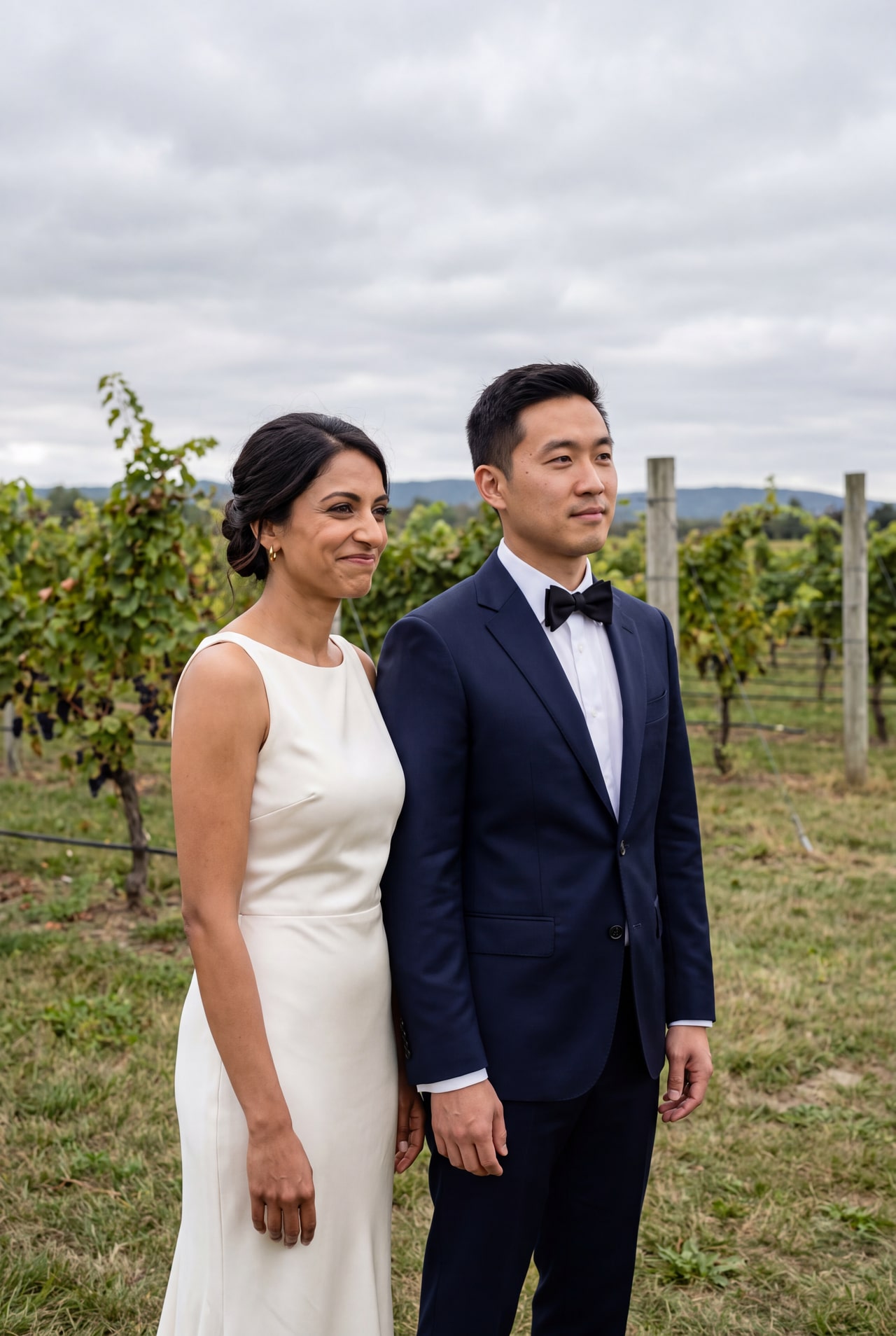 A bride in an ivory dress and groom in a navy suit standing beside vineyard rows